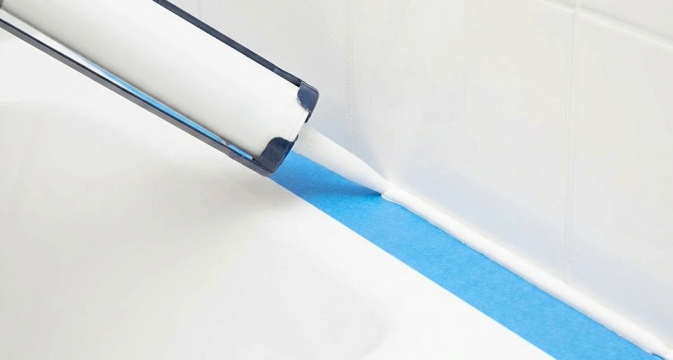 Applying a bead of caulk from a caulk gun along the joint between a bathtub and tile wall, with blue painter's tape on both sides