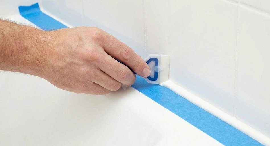 Caulking supplies laid out on a surface: caulk gun loaded with a tube of caulk, blue caulk smoothing tool, roll of blue painter's tape, microfiber cloths, and a utility knife