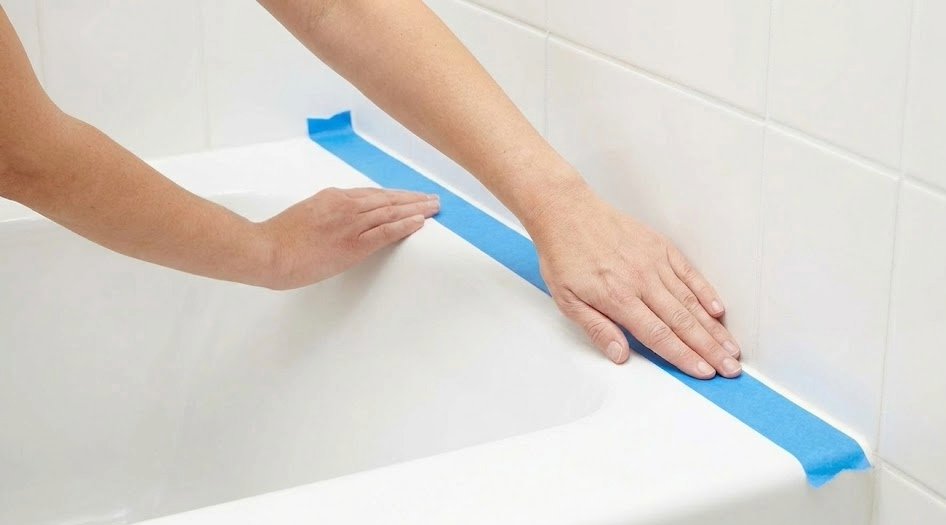 Hands pressing blue painter's tape along the edge of a bathtub where it meets the tile wall, creating a clean straight guide for caulking