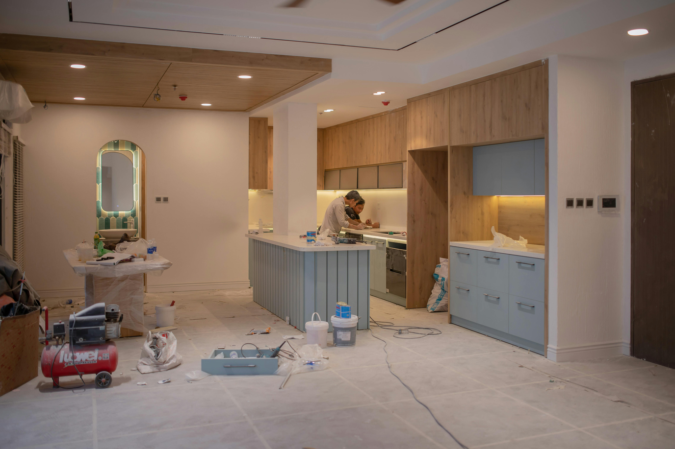 A homeowner rolling soft white paint onto a Shaker-style cabinet door laid on a drop cloth in a sunlit kitchen with freshly painted cabinets in the background