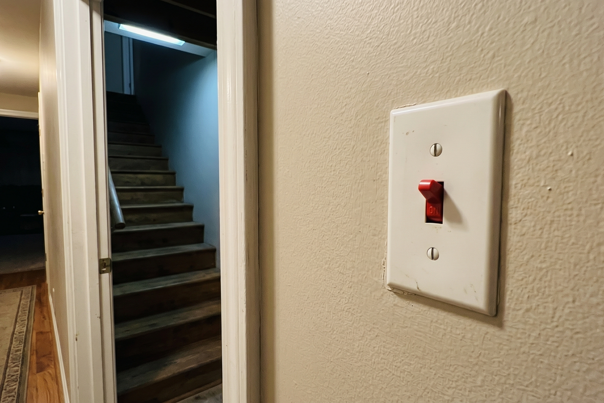 Single red toggle switch in a standard white wall plate mounted at the top of a basement stairwell — a residential HVAC emergency cutoff switch in the ON position